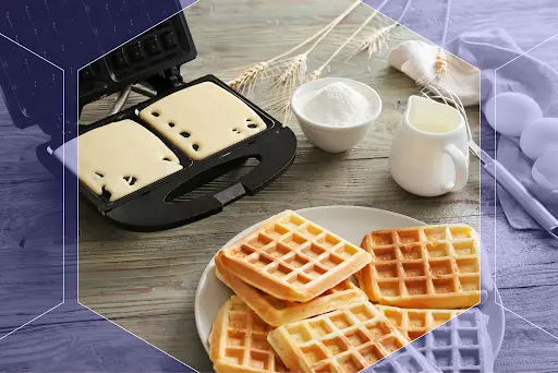 Fresh homemade waffles on a plate beside a waffle maker, flour, milk, and eggs for making waffles from scratch.